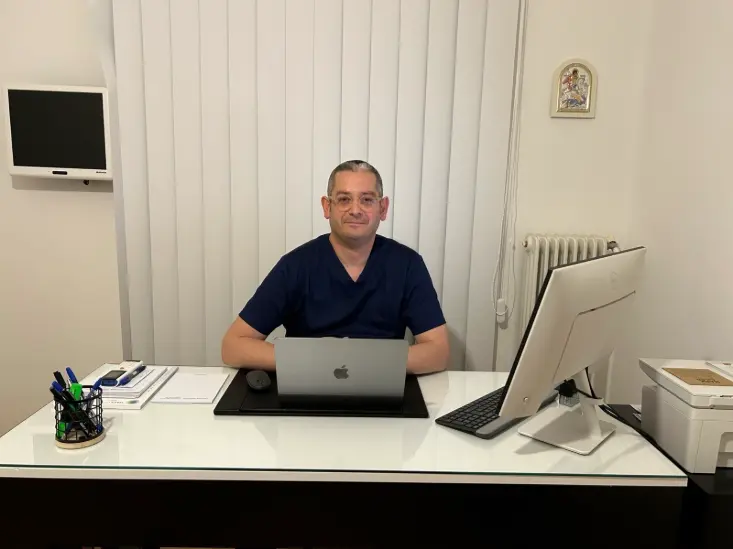 A person in scrubs sits at a desk with a laptop, monitor, and office supplies in a well-lit room.
